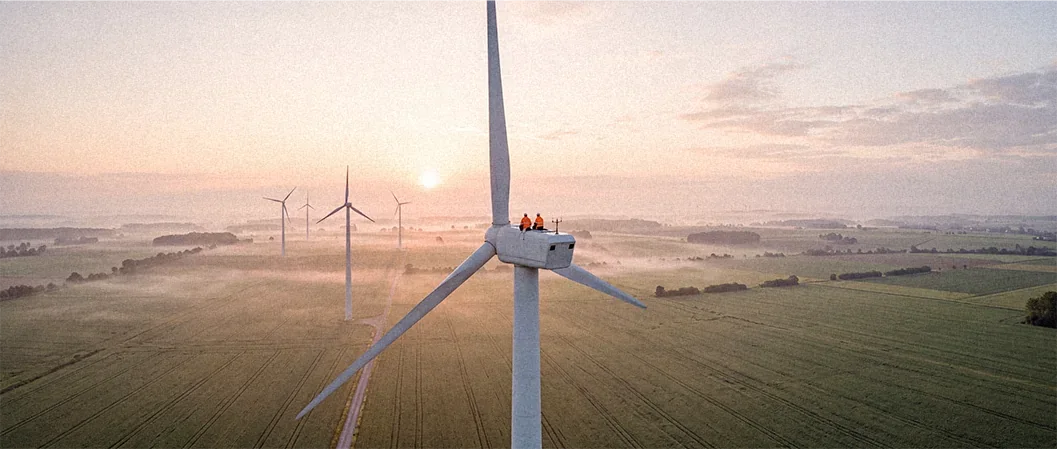 Weite Drohnenaufnahme eines Windrads mit Technikern bei Sonnenaufgang